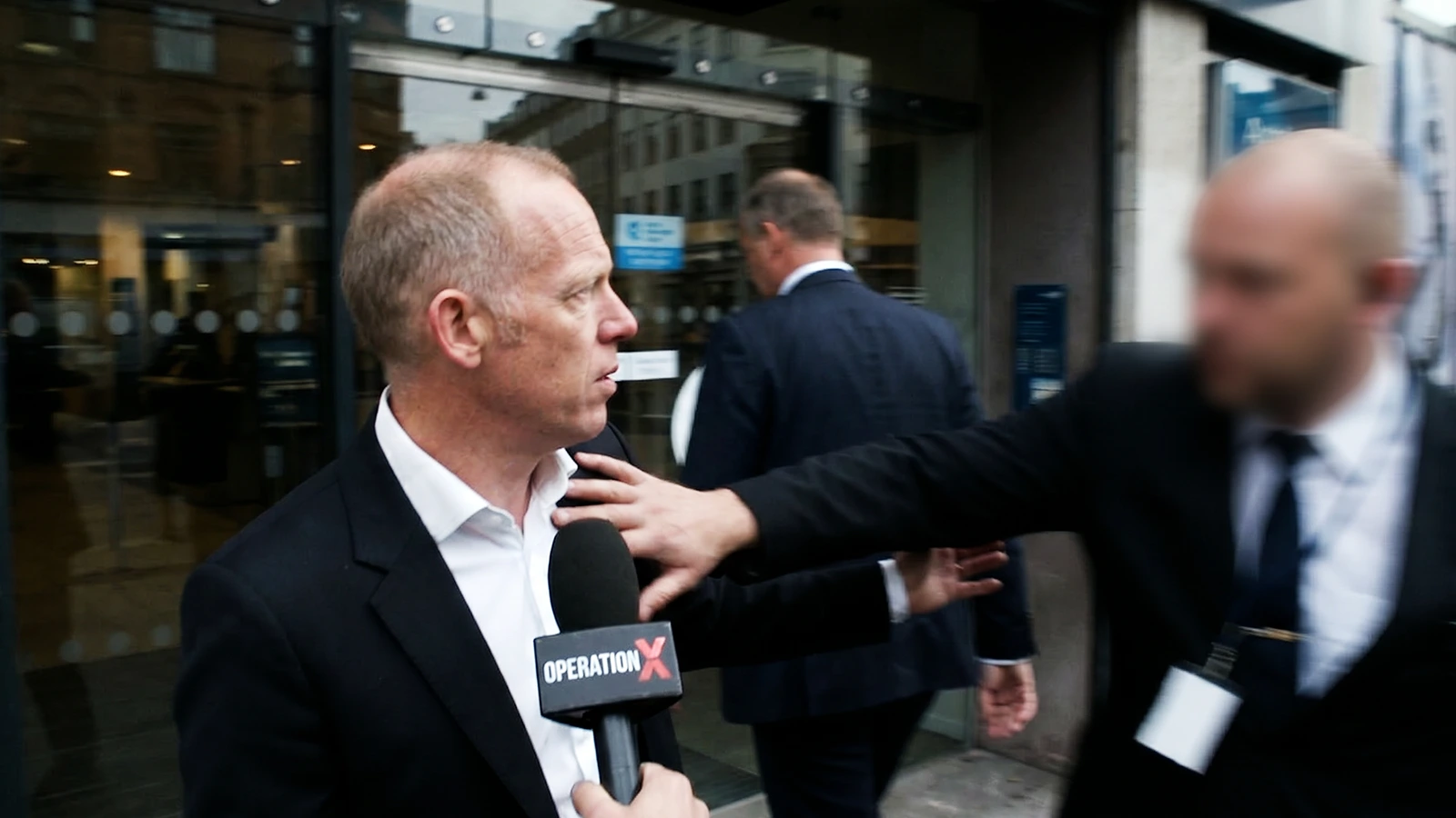 Filialchefen forsvinder ind i Danske Bank, mens vagten blokerer Operation X's hold. Døren lukkes